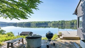 Terraza junto al lago con una parrilla, un banco y una olla grande, con vista a un lago tranquilo y colinas boscosas.