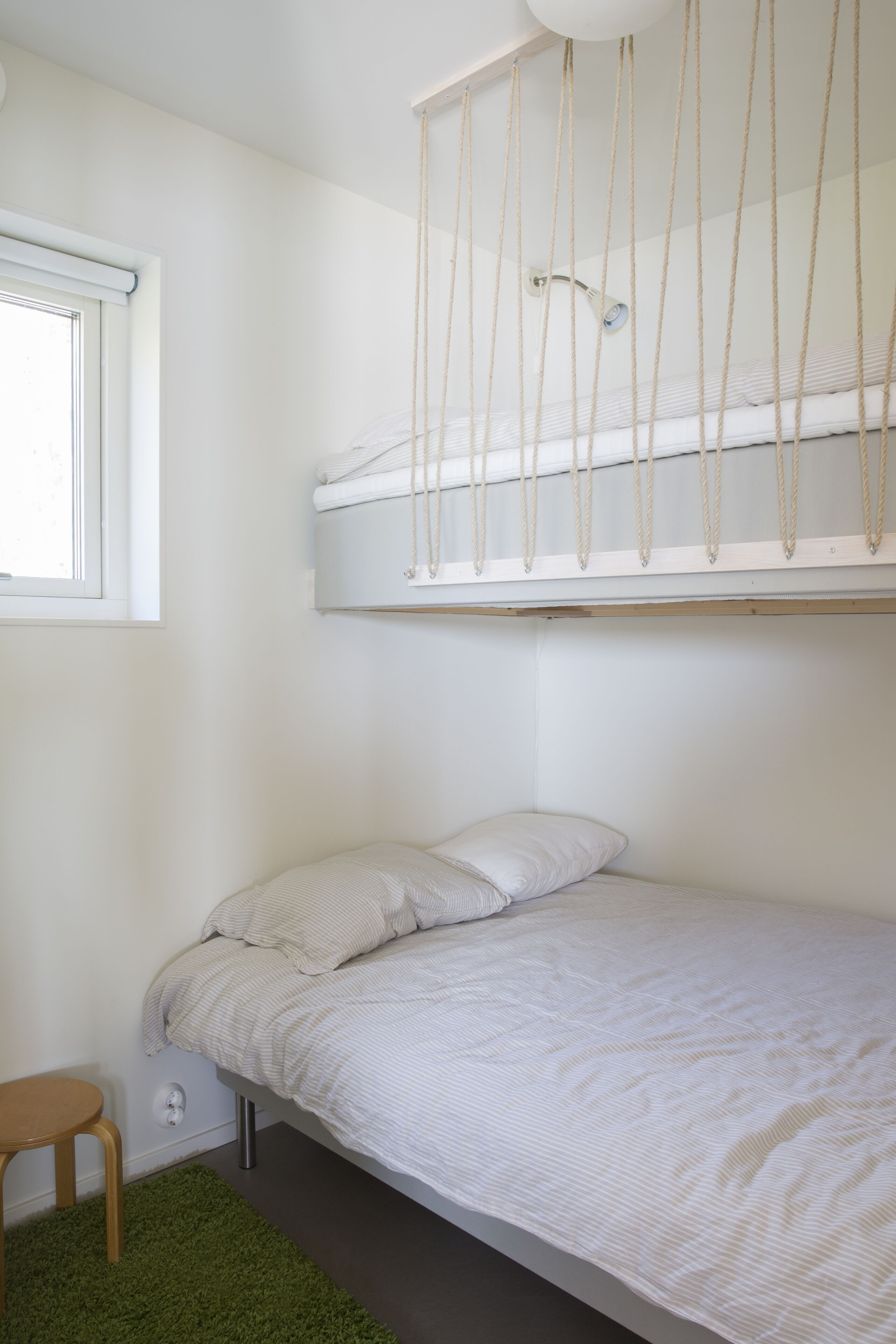 Una cama litera con una hamaca colgante, un taburete de madera pequeño y una ventana con persianas.