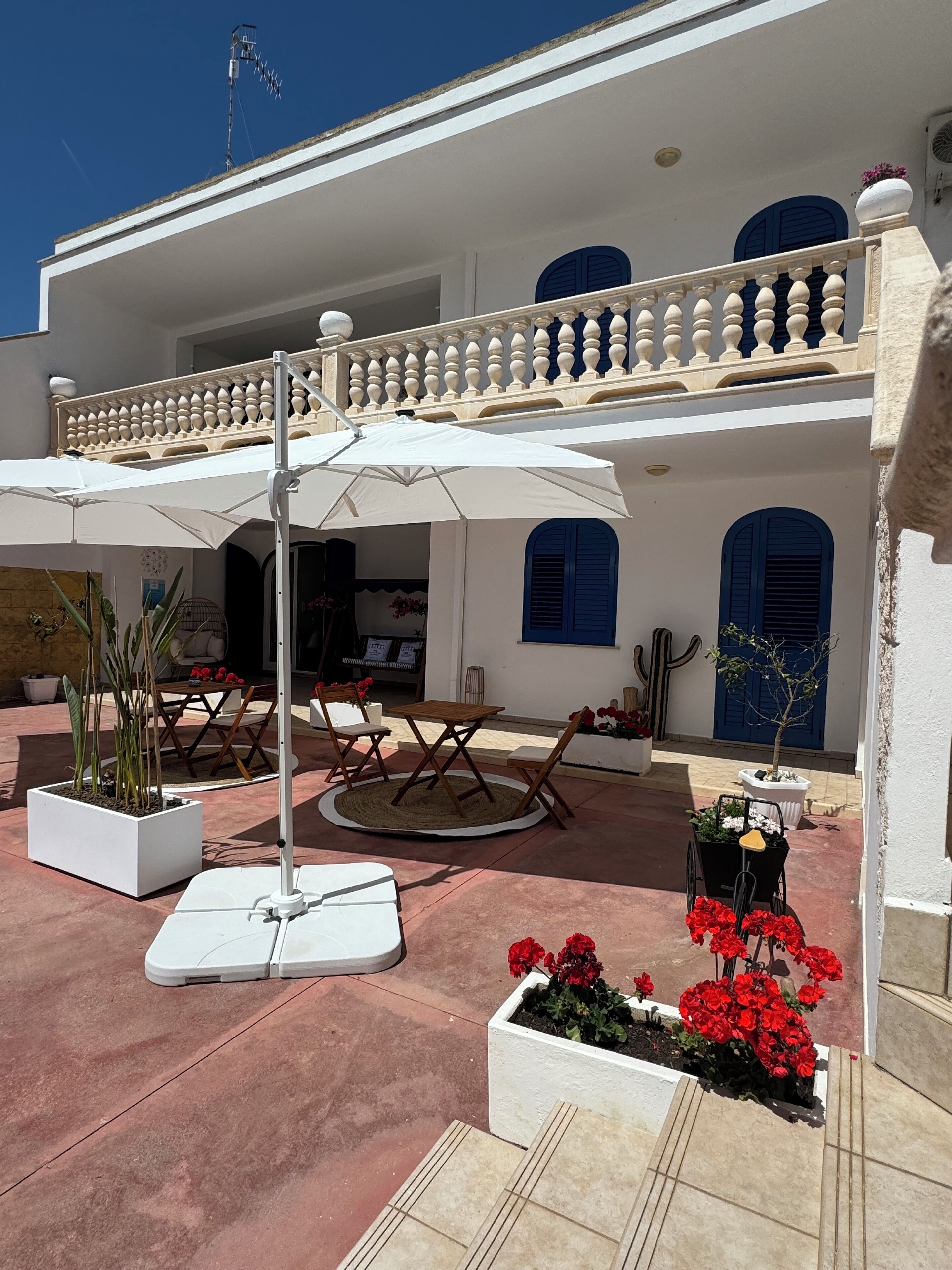 An outdoor patio area with a white umbrella, a round table, and chairs. There are potted plants and a small garden with red flowers.