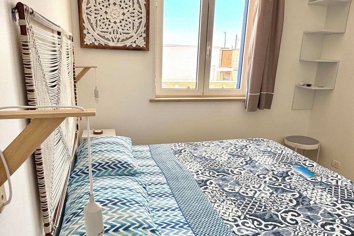 A bedroom with a bed, a bedside table, a wall-mounted shelf, a window with curtains, and a decorative wall piece.