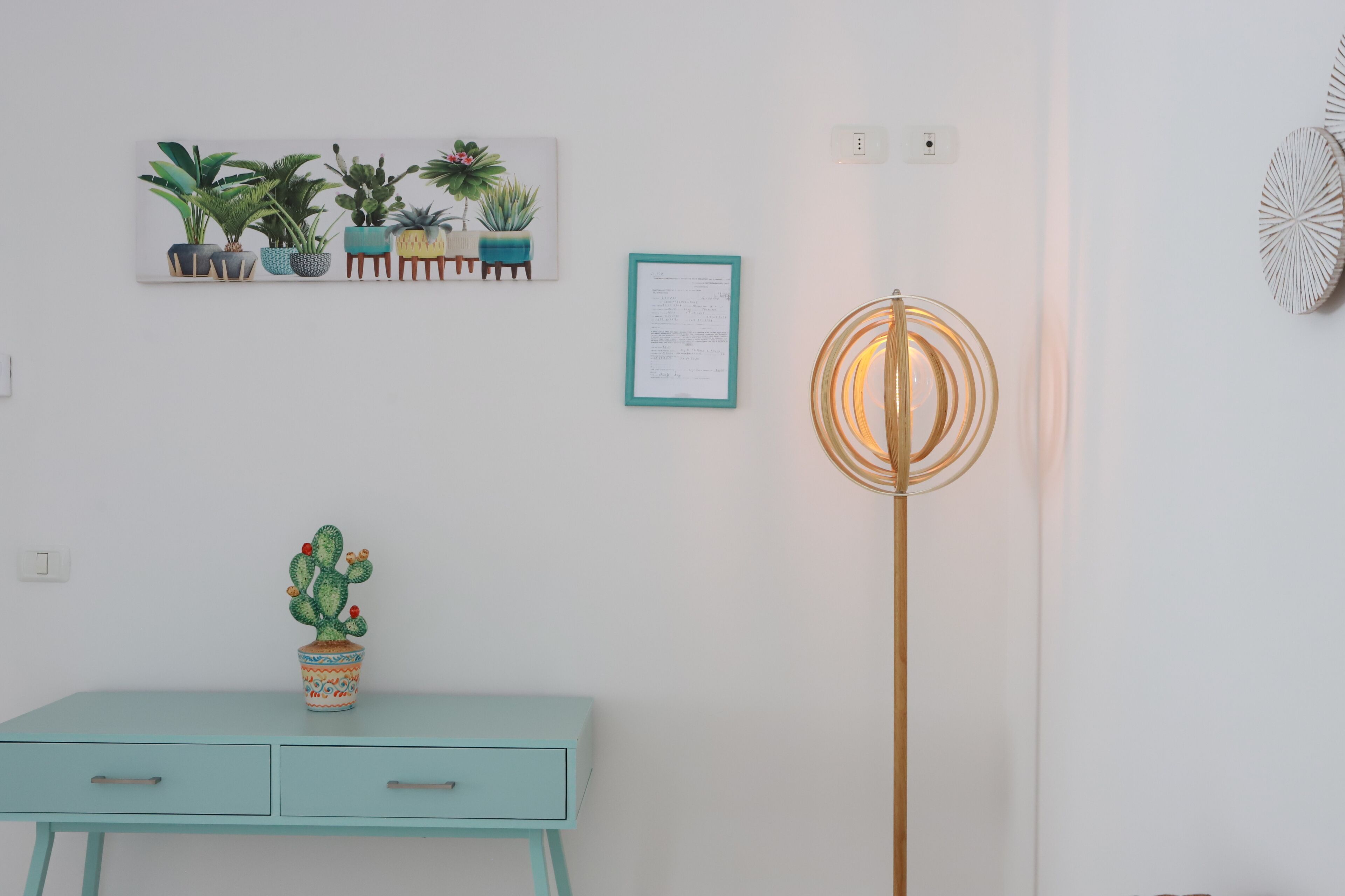 A room with a light blue desk, a cactus in a patterned pot, a framed certificate on the wall, and a tall floor lamp.