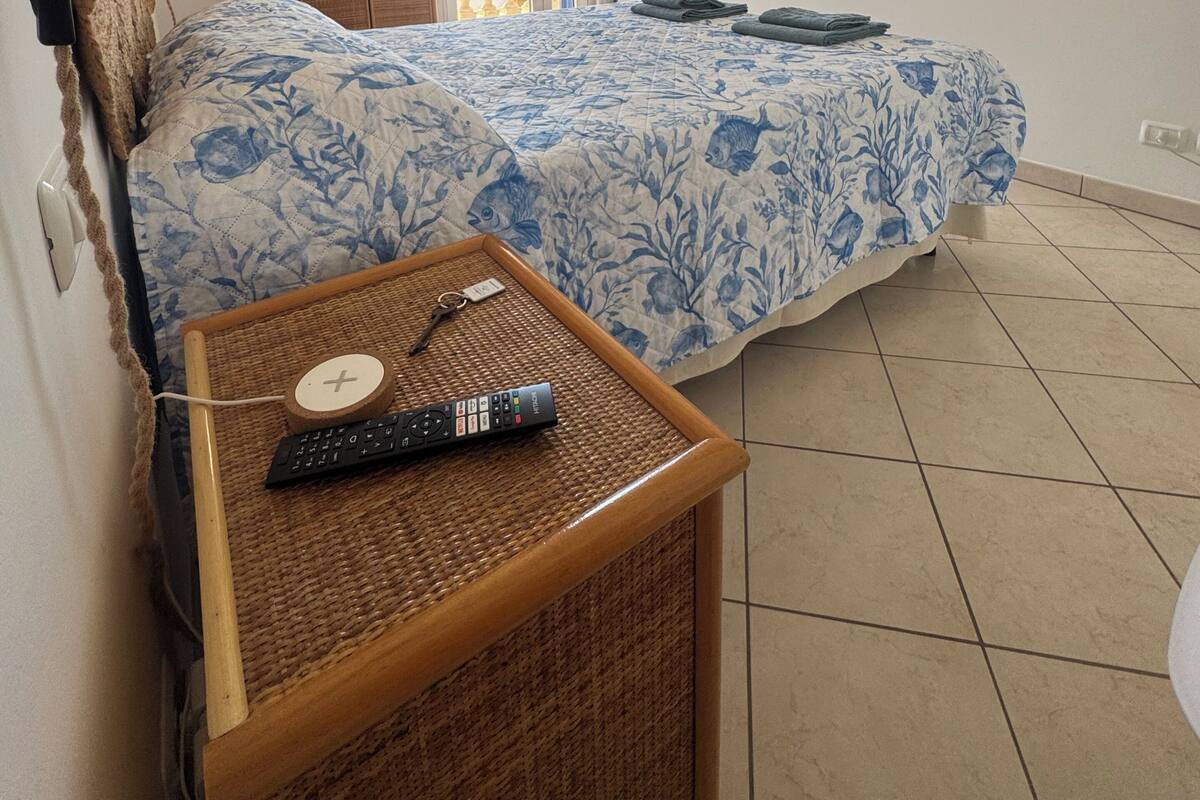 A bedroom with a bed, a bedside table with a remote control, a hanging light fixture, a wardrobe, and a view of the outside.