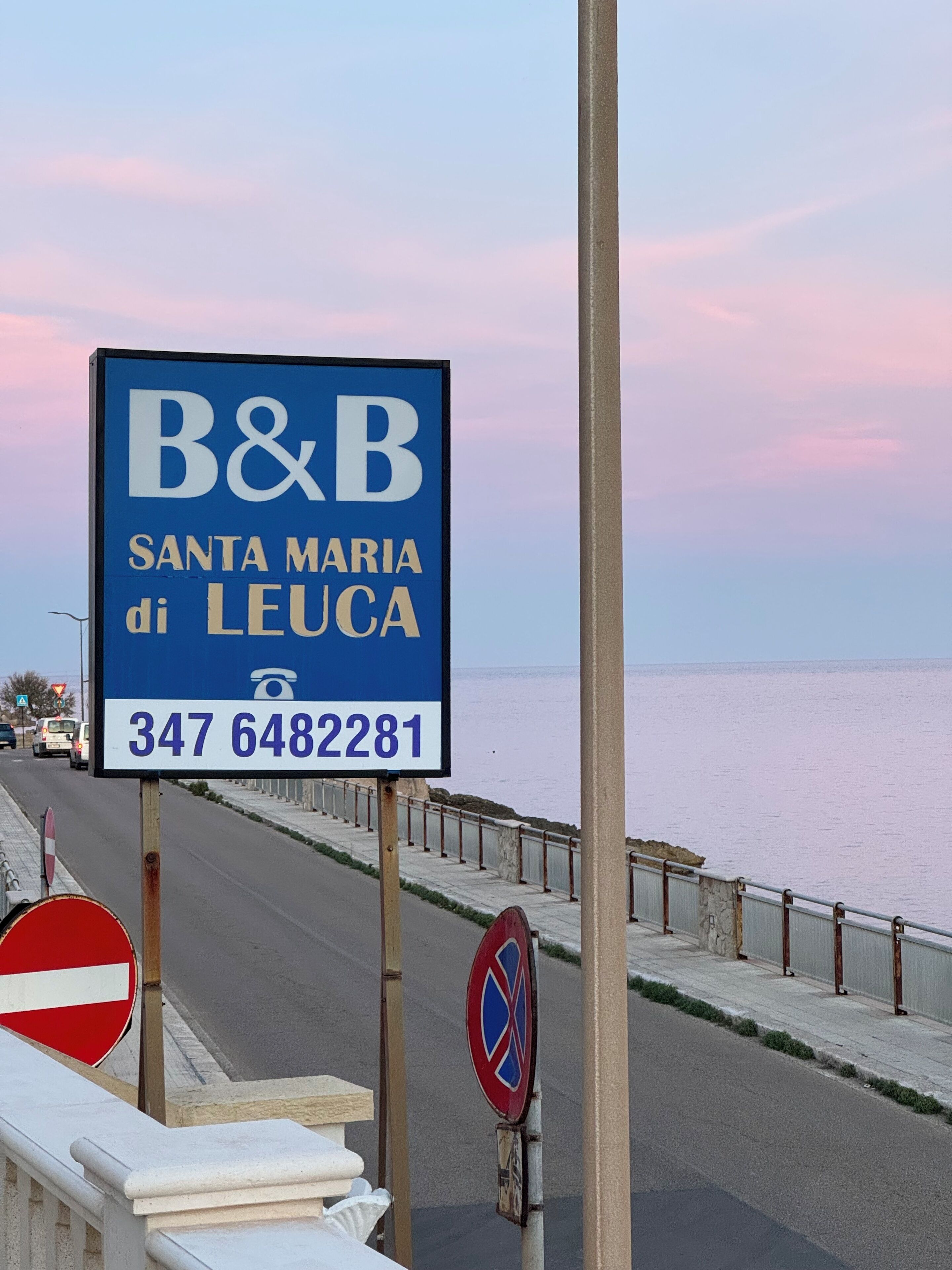 Placa azul com texto branco a anunciar um bed and breakfast em Santa Maria di Leuca, incluindo um número de telefone.