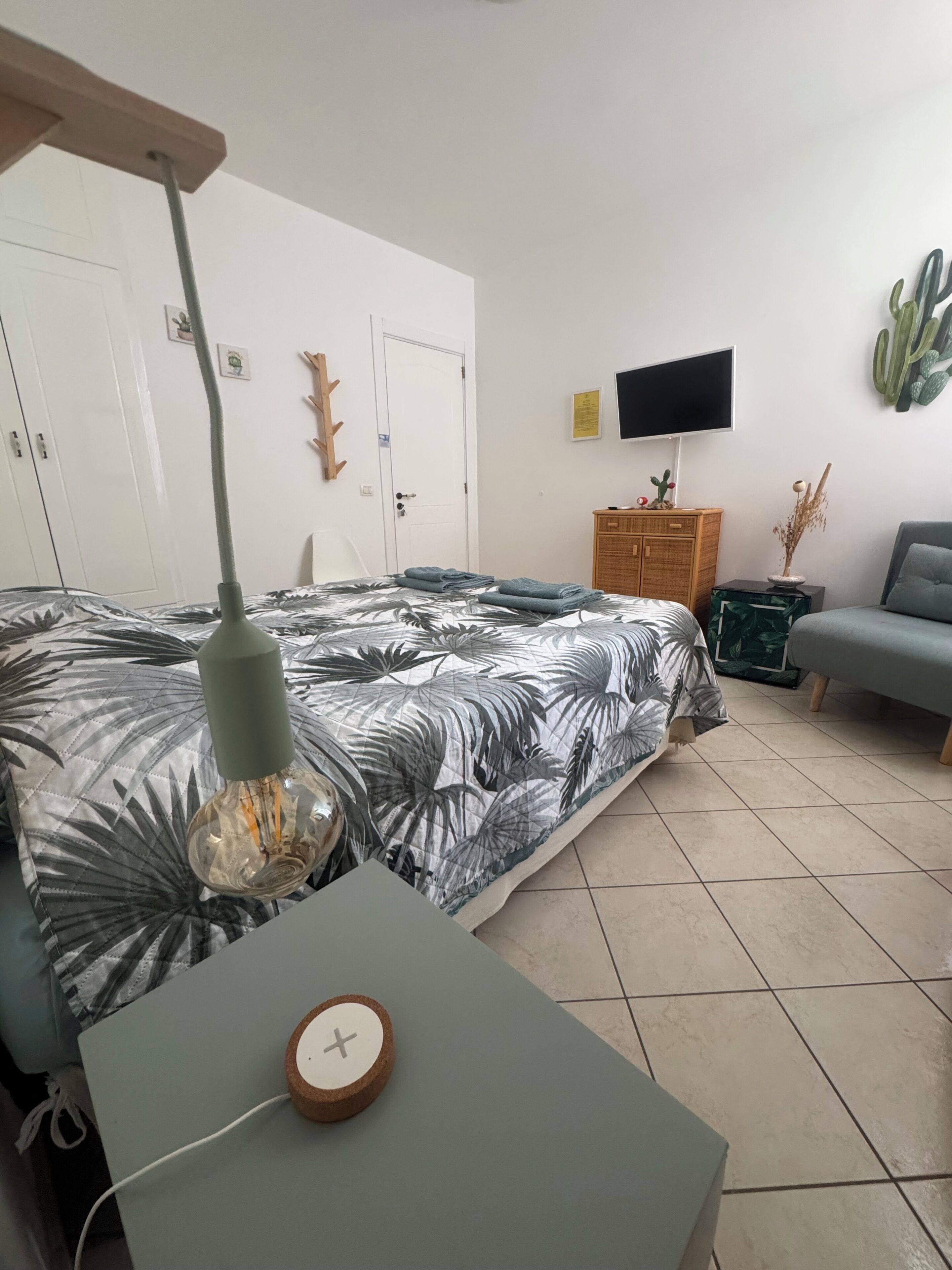 Um quarto com uma cama, uma escrivaninha com abajur, uma televisão, um cacto e um guarda-roupa.