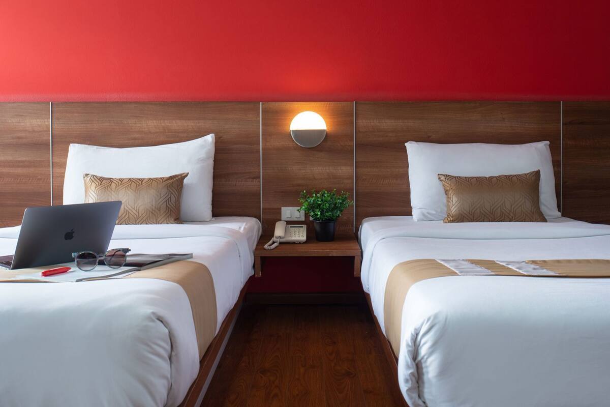 A hotel room with two beds, a desk with a laptop, a phone, and a plant.