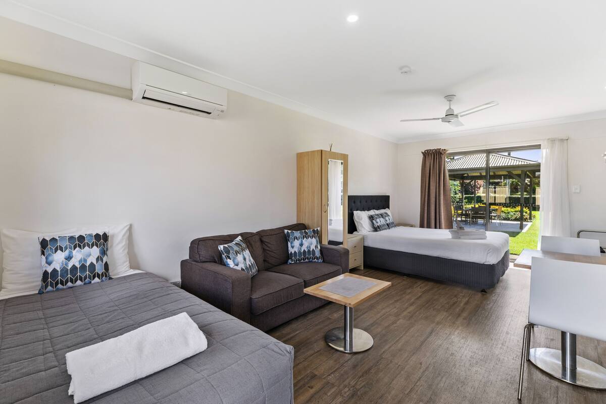 A modern hotel room with two beds, a sofa, a small table, and a balcony with a view of a garden.