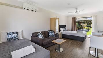 A modern hotel room with two beds, a sofa, a small table, and a balcony with a view of a garden.