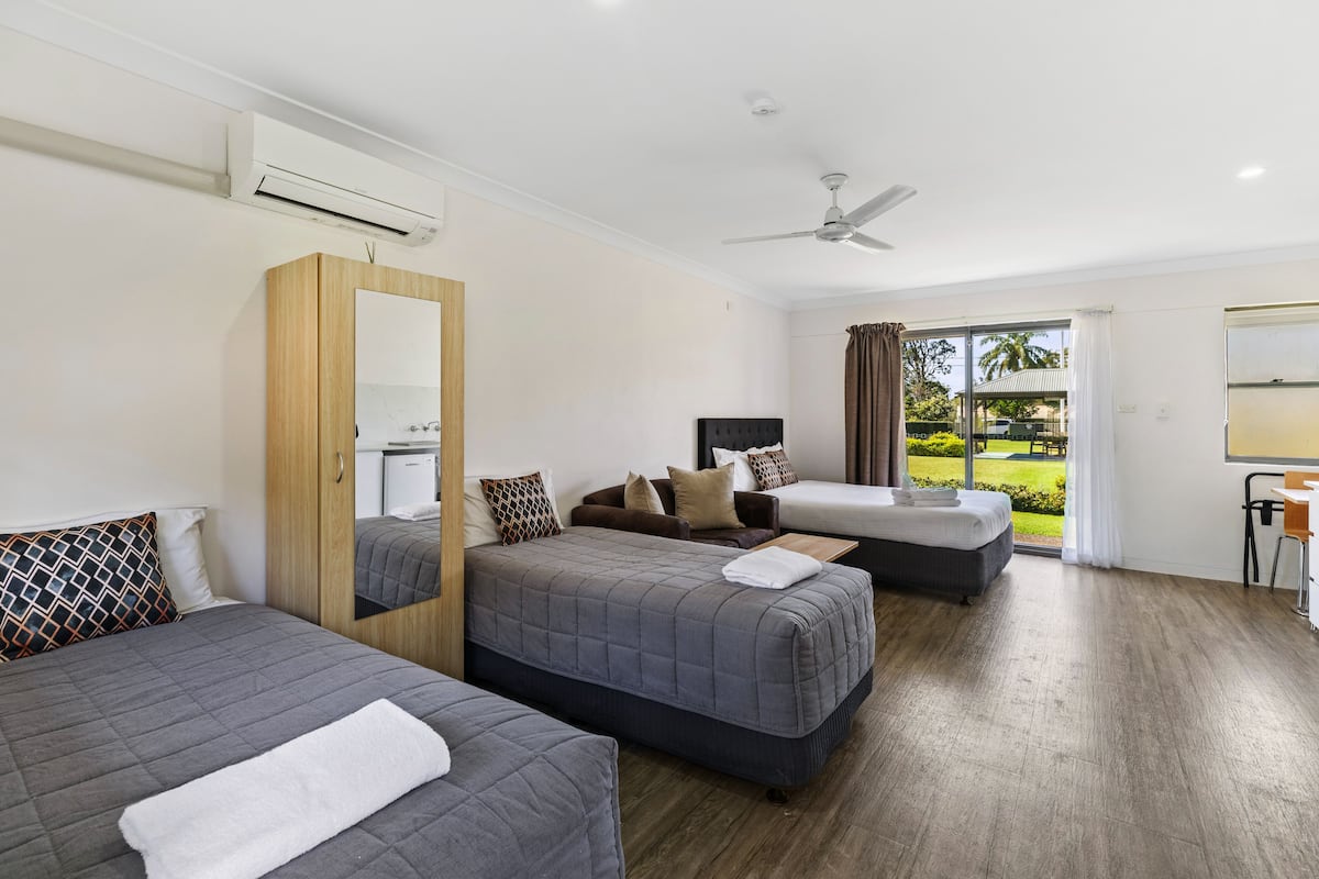 A hotel room with three beds, a ceiling fan, an air conditioning unit, a large window with curtains, and a view of the outdoors.