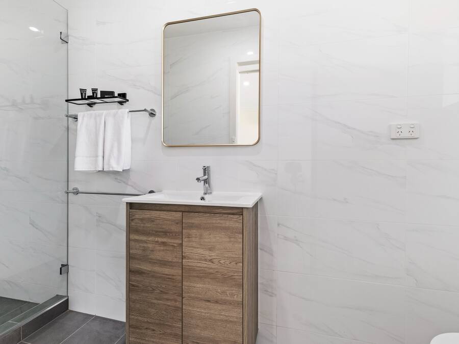 A modern bathroom with a marble wall, a wooden vanity, a white sink, and a large mirror.