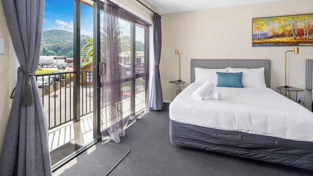 A hotel room with a large bed, a painting on the wall, and a balcony with a view of a mountain and a parking area. - ASURE Rotorua International Motor Inn (Rotorua)