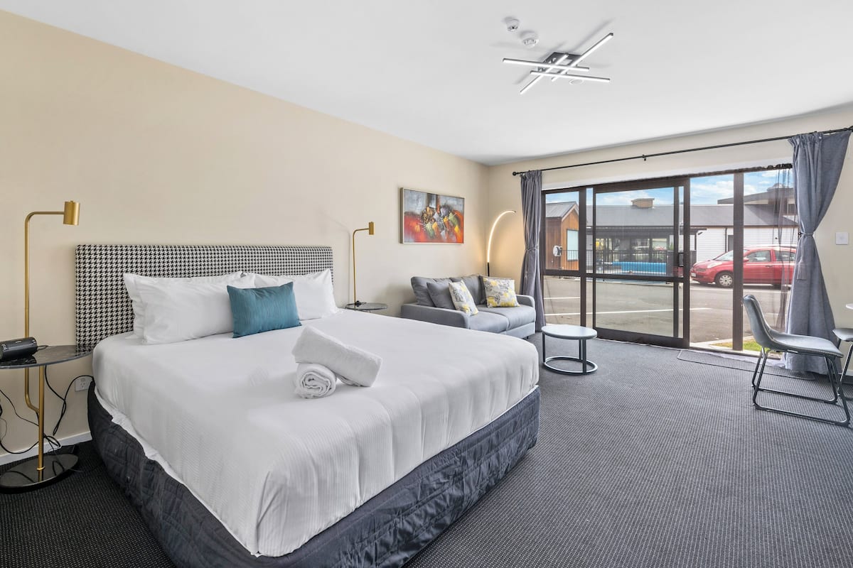 A hotel room with a large bed, a sofa, a small table, and a view of the street through a large window.
