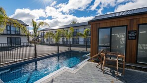 A swimming pool with a wooden deck and a bench, surrounded by a fence and palm trees. - ASURE Rotorua International Motor Inn (Rotorua)