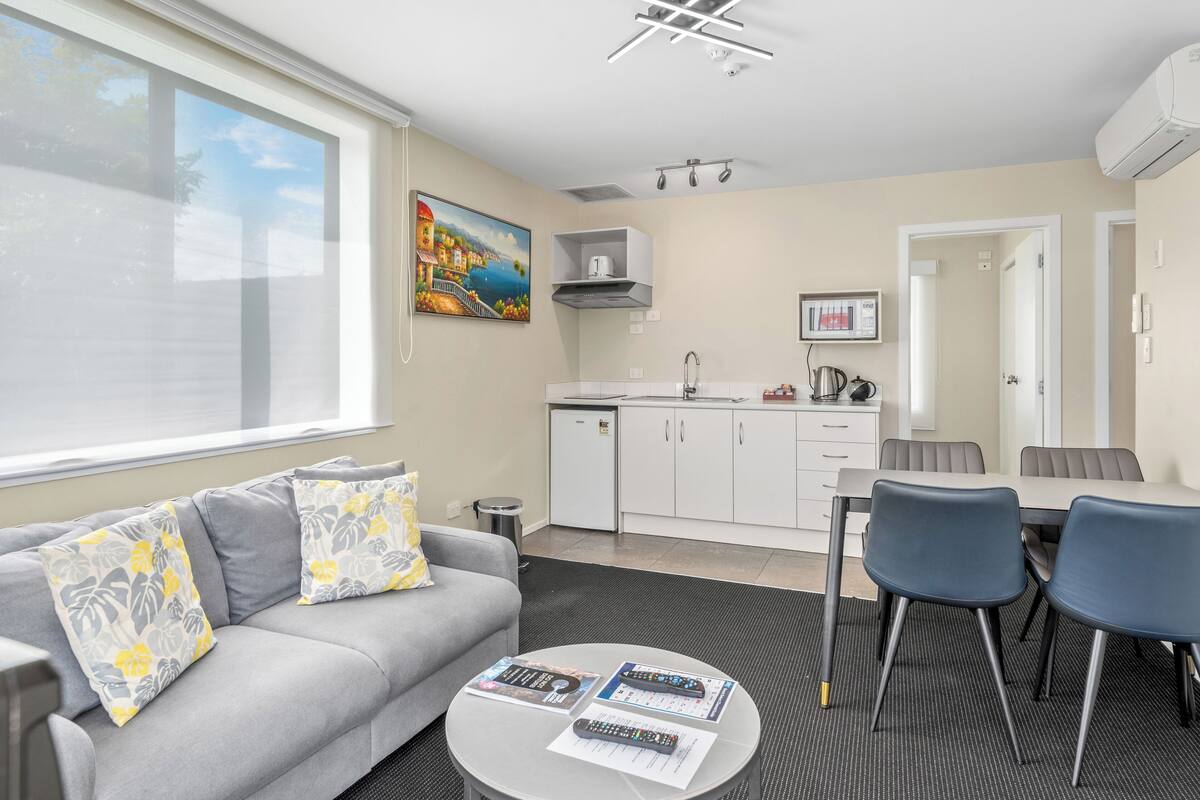 A modern living room with a grey sofa, a coffee table with remote controls, a dining area with a table and chairs, and a kitchen with white cabinets.