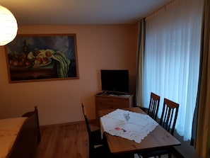Un coin repas comprenant une table, des chaises, un tableau, un téléviseur et une fenêtre avec des rideaux.