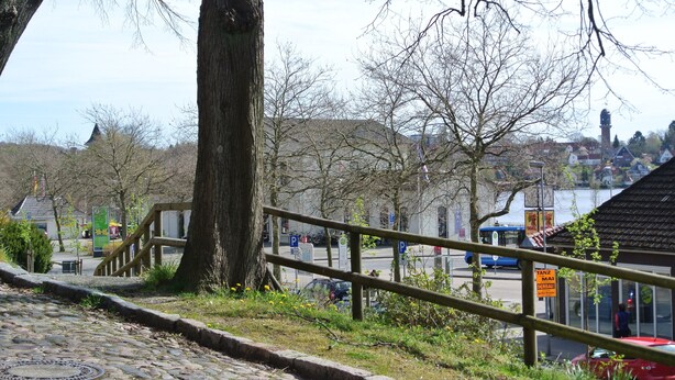 Un camino adoquinado que conduce a una escalera de madera, una barandilla metálica y vistas de un pueblo con edificios y una masa de agua al fondo.