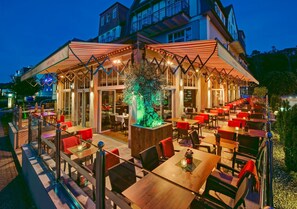 Een openluchtrestaurant met tafels en stoelen, een boom in het midden en een gebouw op de achtergrond.
