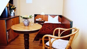 Quarto de hotel com mesa de madeira, cadeira de vime, cama com manta vermelha, mesa de cabeceira com abajur e um vaso com flores.