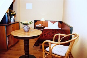 Hotelkamer met een houten tafel, een rieten stoel, een bed met rode deken, een nachtkastje met lamp en een vaas met bloemen.