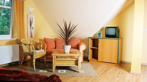 Uma sala de estar aconchegante com um sofá, duas poltronas, uma mesa de centro, uma televisão sobre um móvel e uma janela com cortinas.