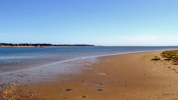 Ein weiter Sandstrand mit ruhiger See, blauem Himmel und einer fernen Küstenlinie mit Bäumen und Gebäuden.