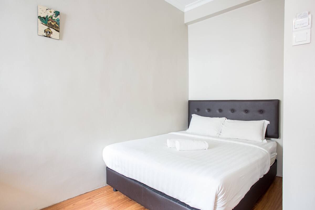 Ein ordentlich bezogenes Bett mit weißen Leinen und einem dunklen Kopfteil in einem schlichten Zimmer mit weißen Wänden und einem kleinen Bilderrahmen.