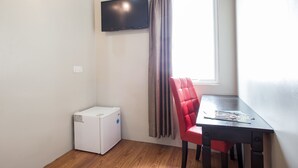 A room with a flat-screen TV mounted on the wall, a window with curtains, a compact white appliance, a red chair, and a small table with magazines. - Moment Hotel (Petaling Jaya)