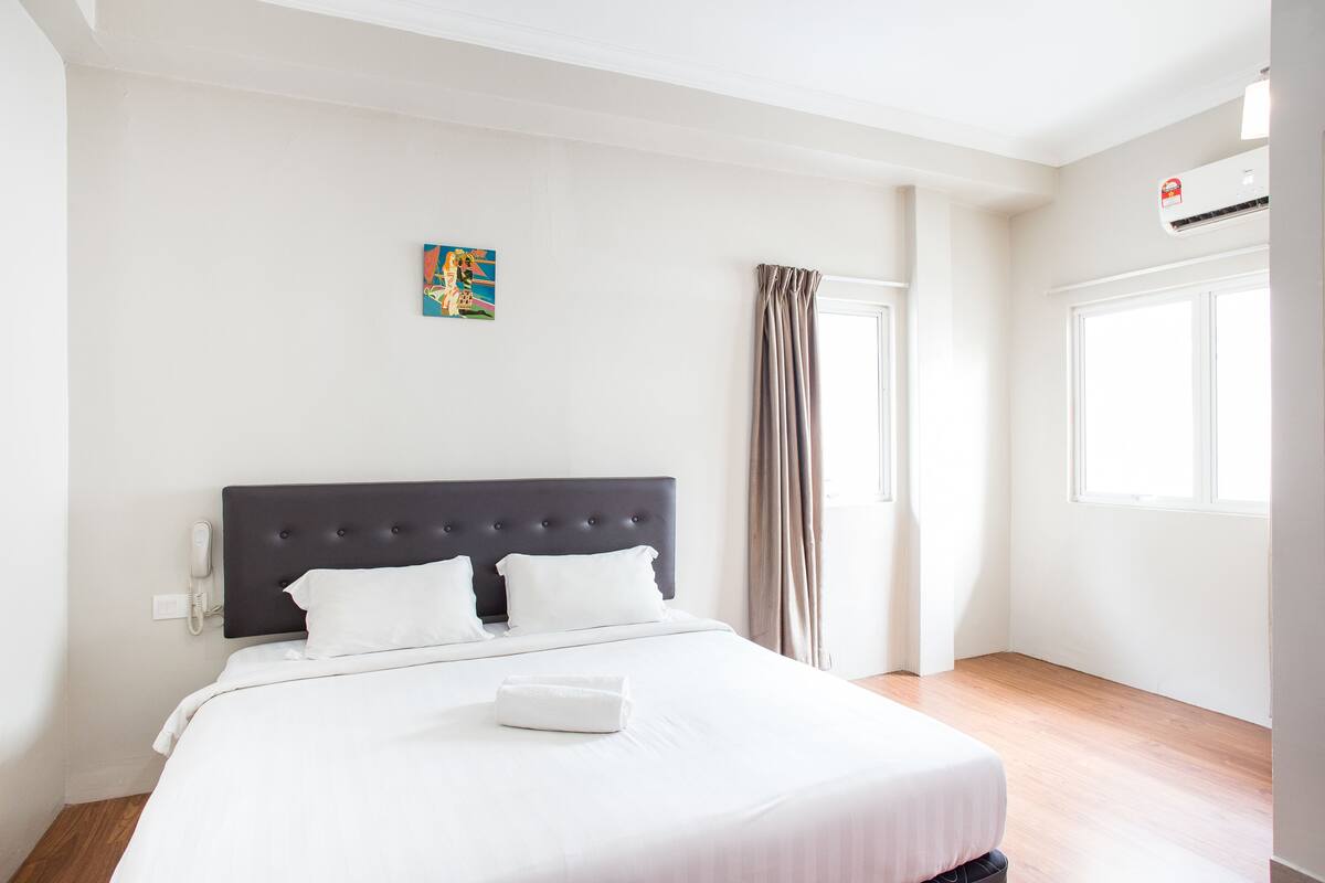 Ein Hotelzimmer mit einem großen Bett, einer Kopfteil, einem Nachttischtelefon, einem Fenster mit Vorhängen und einer Klimaanlage.