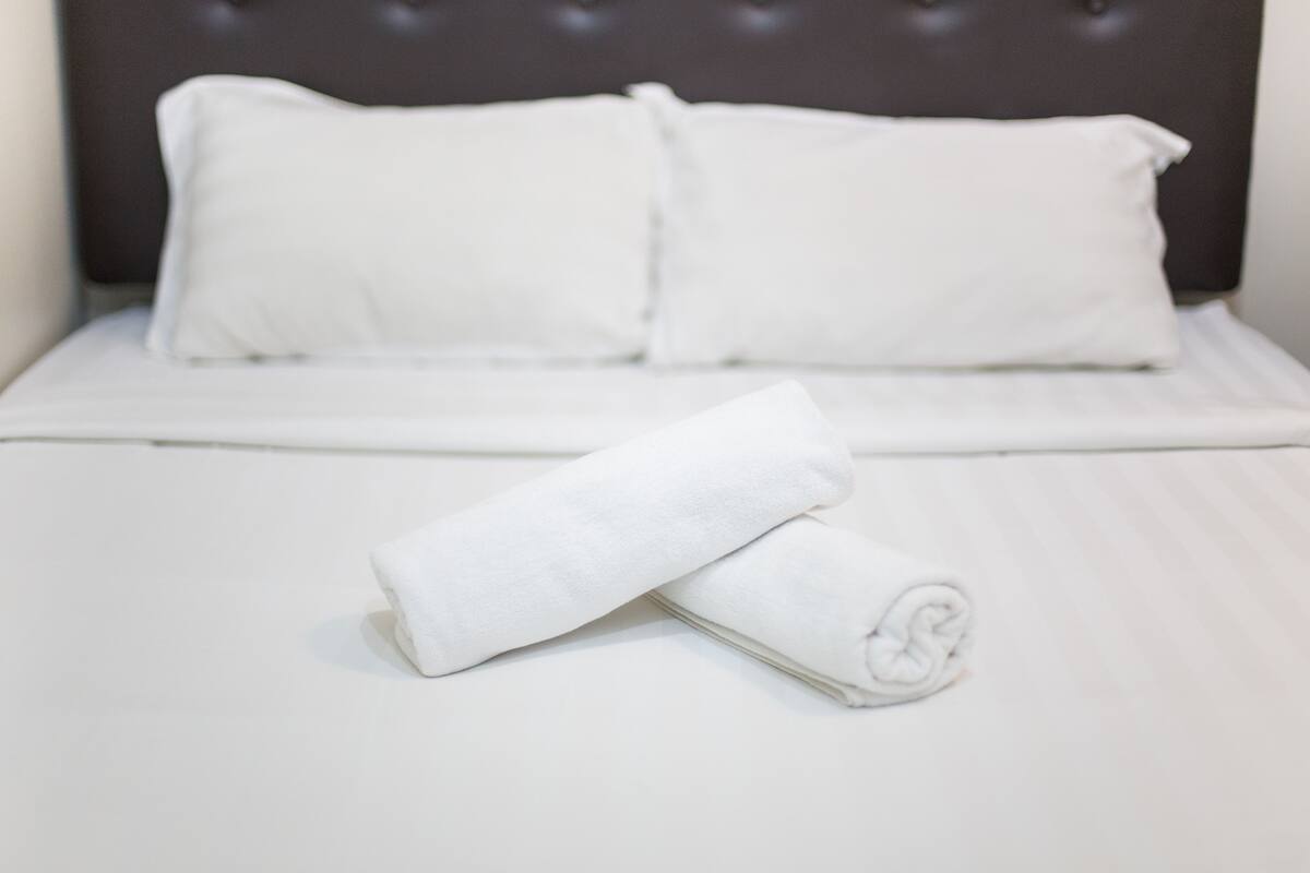 Ein ordentlich bezogenes Bett mit weißen Leinen und Kissen, ein aufgerolltes weißes Handtuch auf dem Bett und ein dunkles Kopfteil.