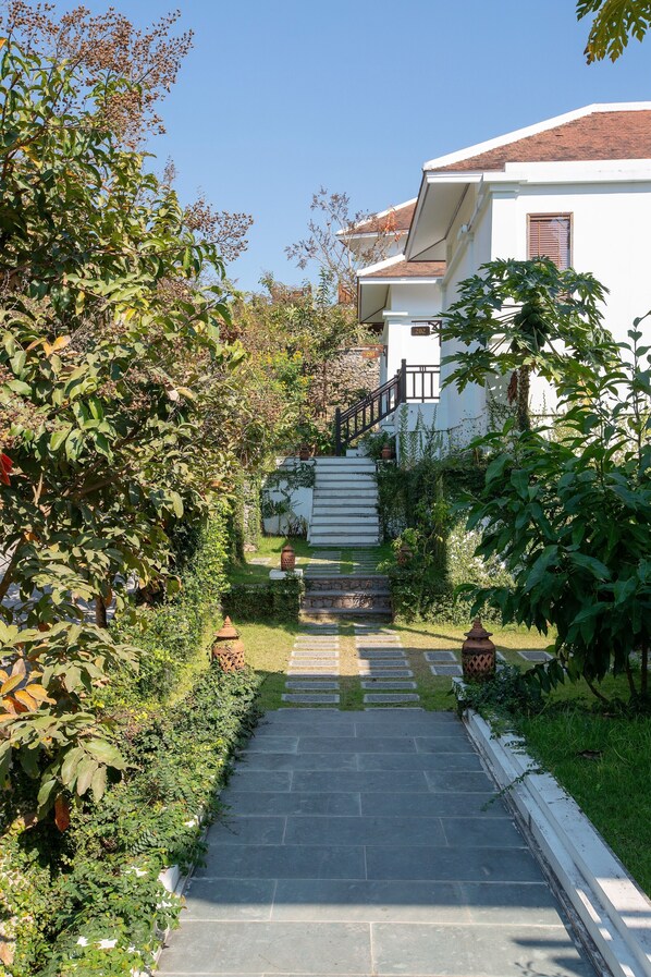 A stone pathway leading to a white building with a red roof, surrounded by lush greenery and a small garden.