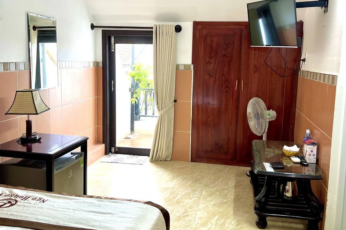 A hotel room with a bed, a desk with a lamp, a TV mounted on the wall, a fan, and a small table with items on it.