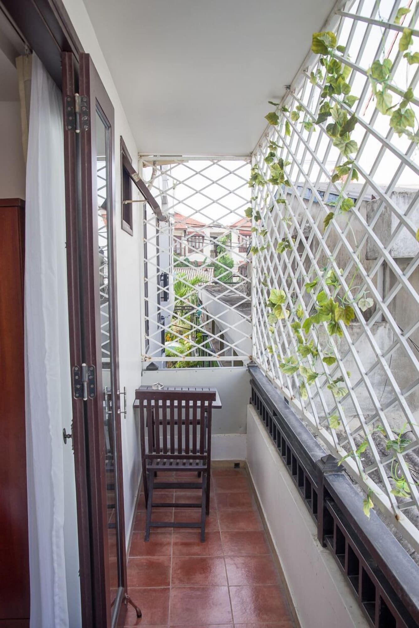 A narrow balcony with a wooden chair, a white lattice fence, and a view of buildings in the background.