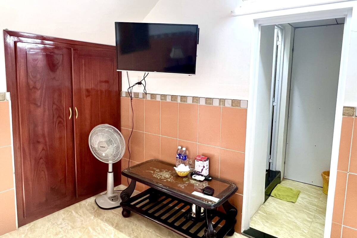 A room with a wooden wardrobe, a standing fan, a table with items, and a TV mounted on the wall.