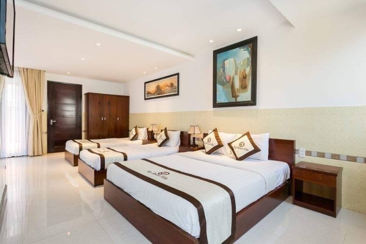 A hotel room with four beds arranged in a row, a wooden wardrobe, a painting on the wall, and a small wooden table beside the beds.