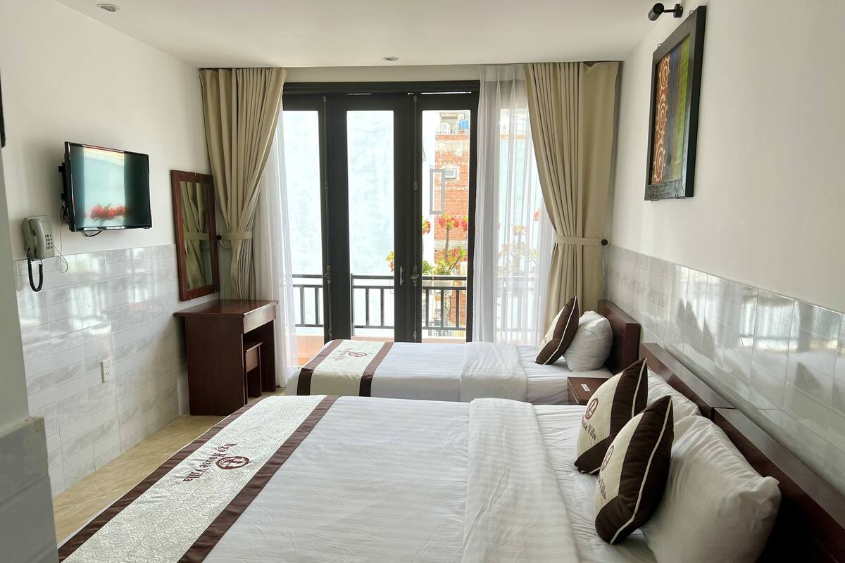 A hotel room with two beds, a TV, a phone, and a balcony with a view of buildings and plants.
