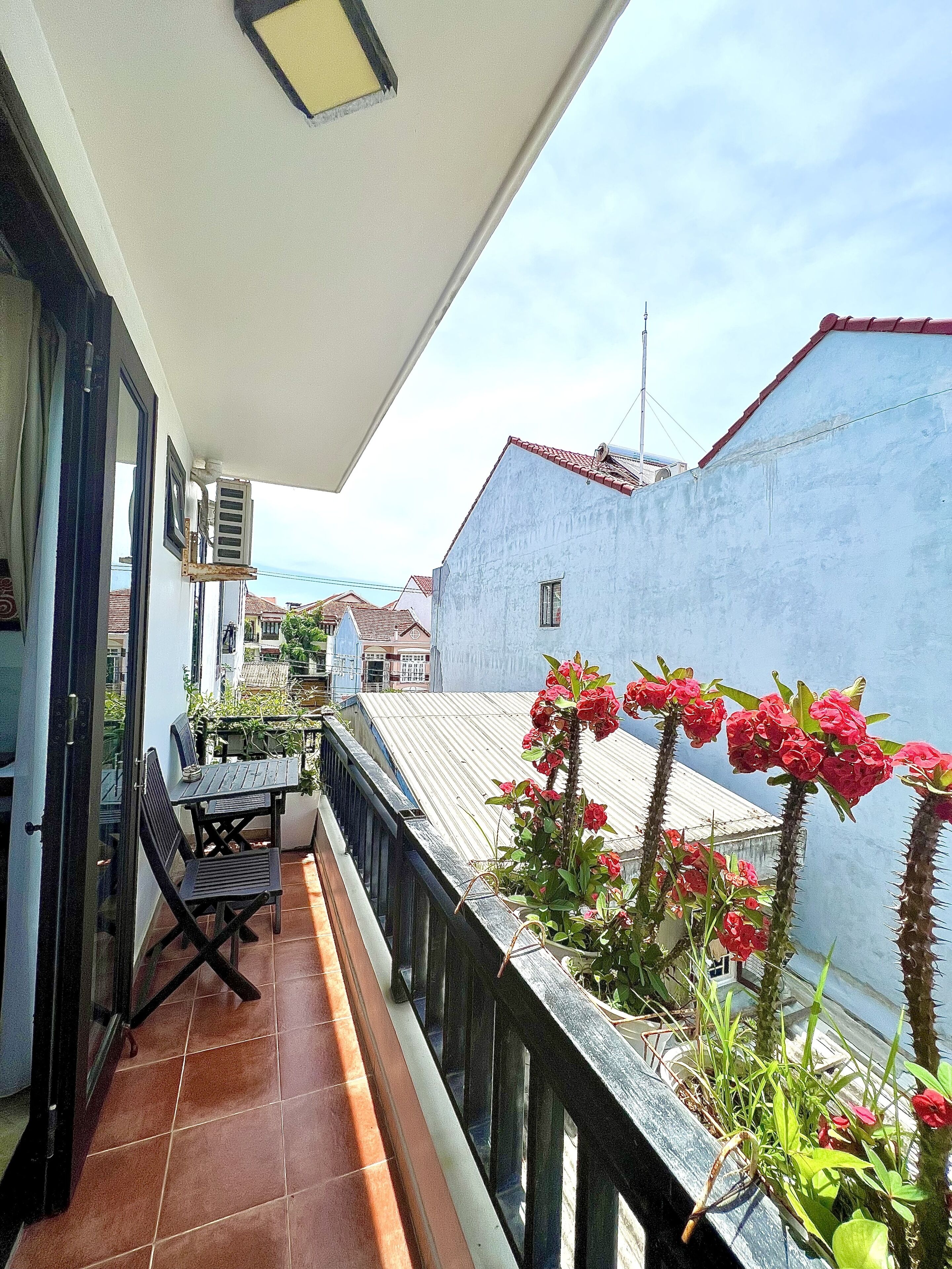 A balcony with a table and chairs, a potted plant with red flowers, and a view of neighboring buildings.