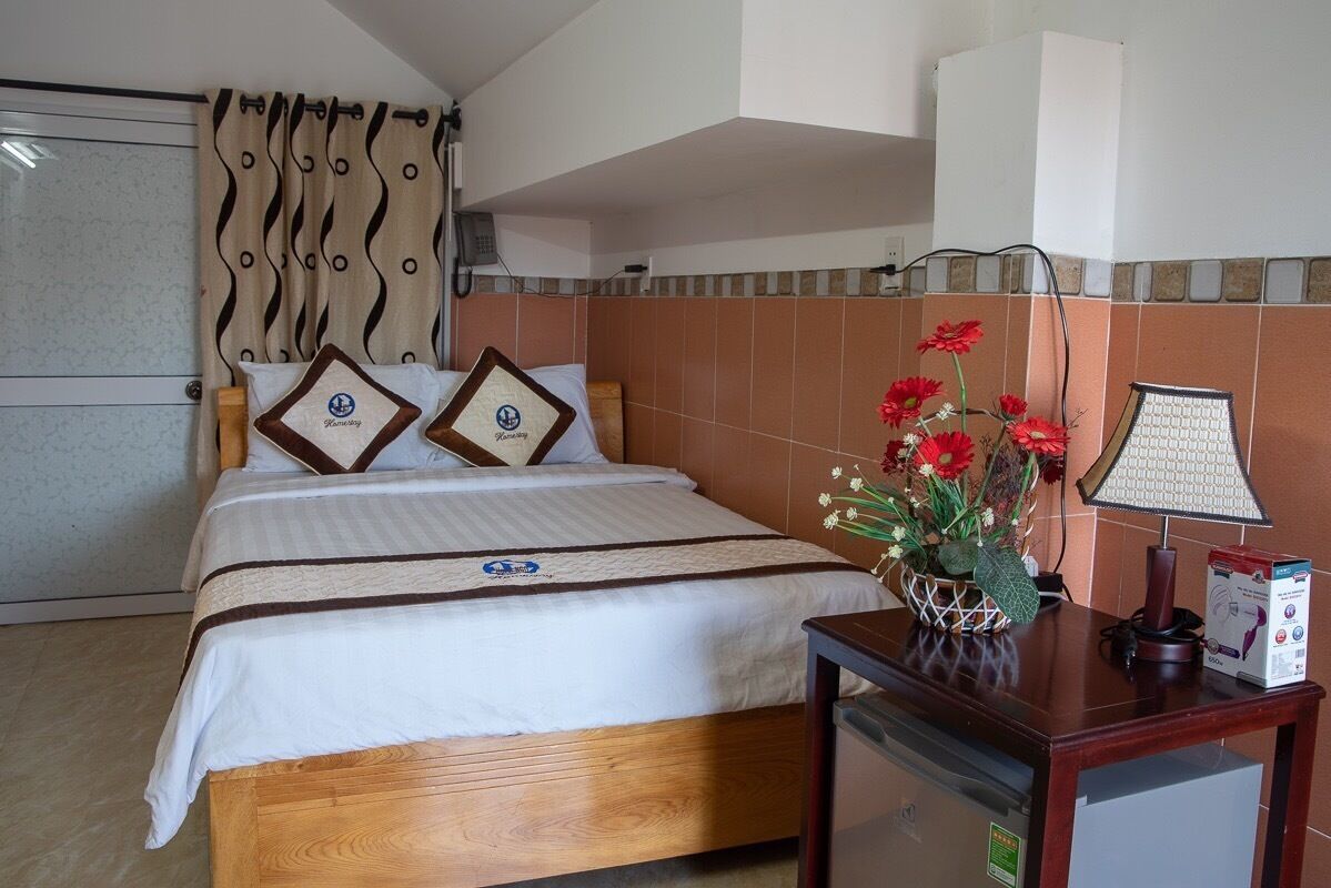 A hotel room with a bed, a bedside table with a lamp, a vase with flowers, and a small cabinet.