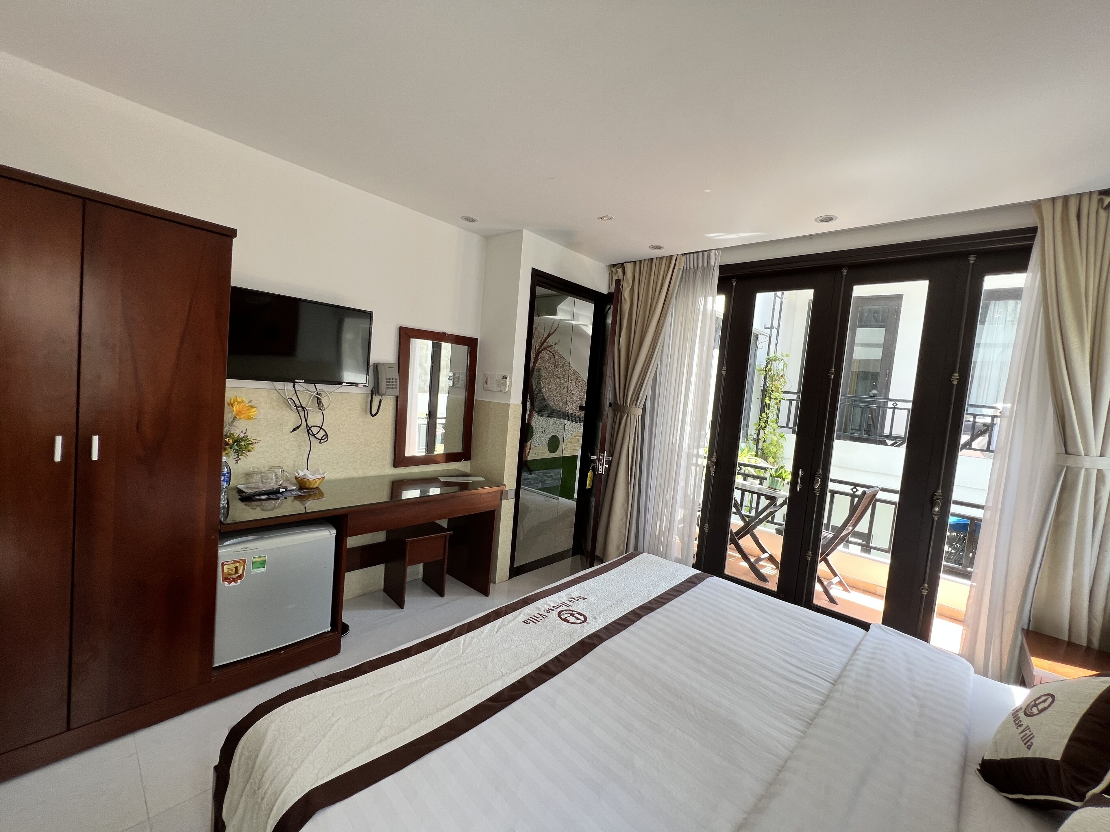 Habitación de hotel con cama, escritorio, armario, televisor y balcón con cortinas.