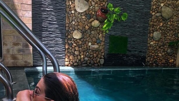 Eine Person entspannt sich in einem Schwimmbecken mit klarem Wasser, umgeben von einer Steinmauer und einem Metallgeländer.