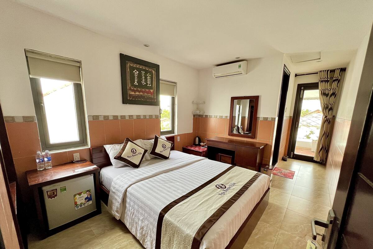 A hotel room with a bed, bedside tables, a desk, a mirror, and an air conditioning unit.