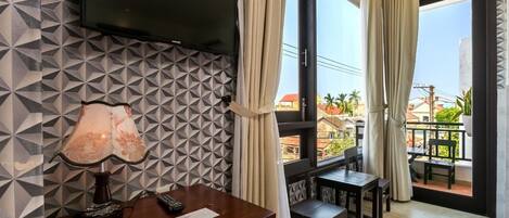 A hotel room with a TV mounted on the wall, a desk with a lamp, and a balcony with a table and chairs.