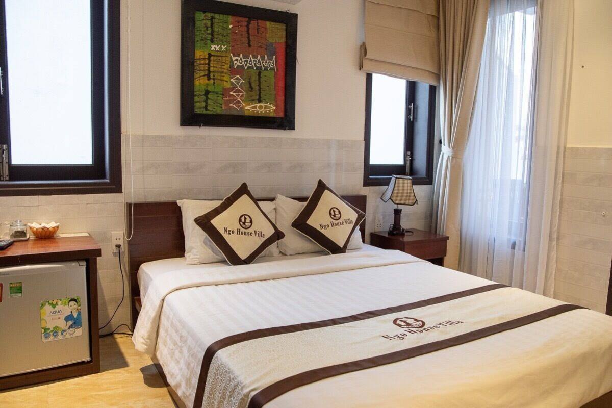 A hotel room with a bed, a small desk with a bowl, a refrigerator, and a window with curtains.