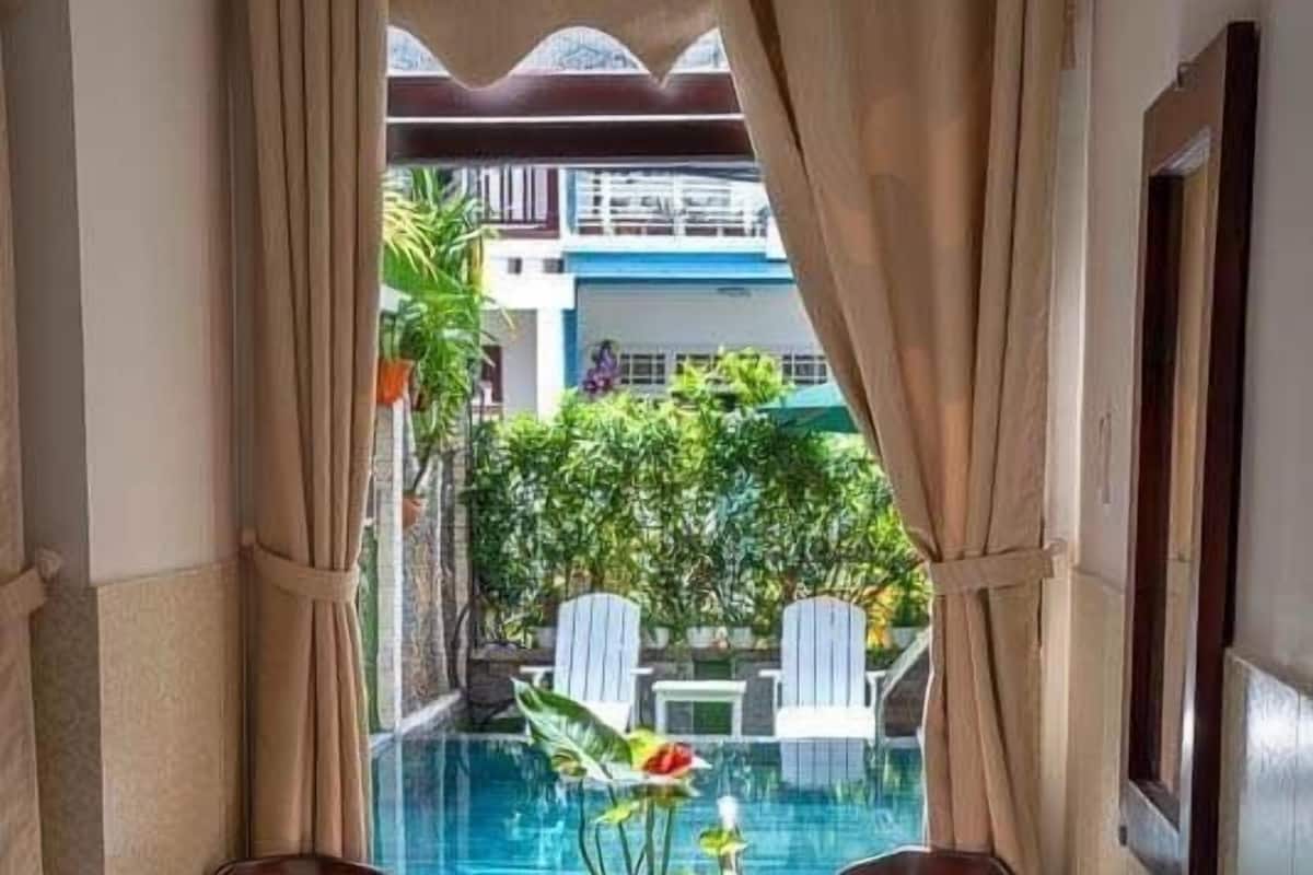 A corner room with a view of a swimming pool, two wooden chairs, a small table, and a potted plant.