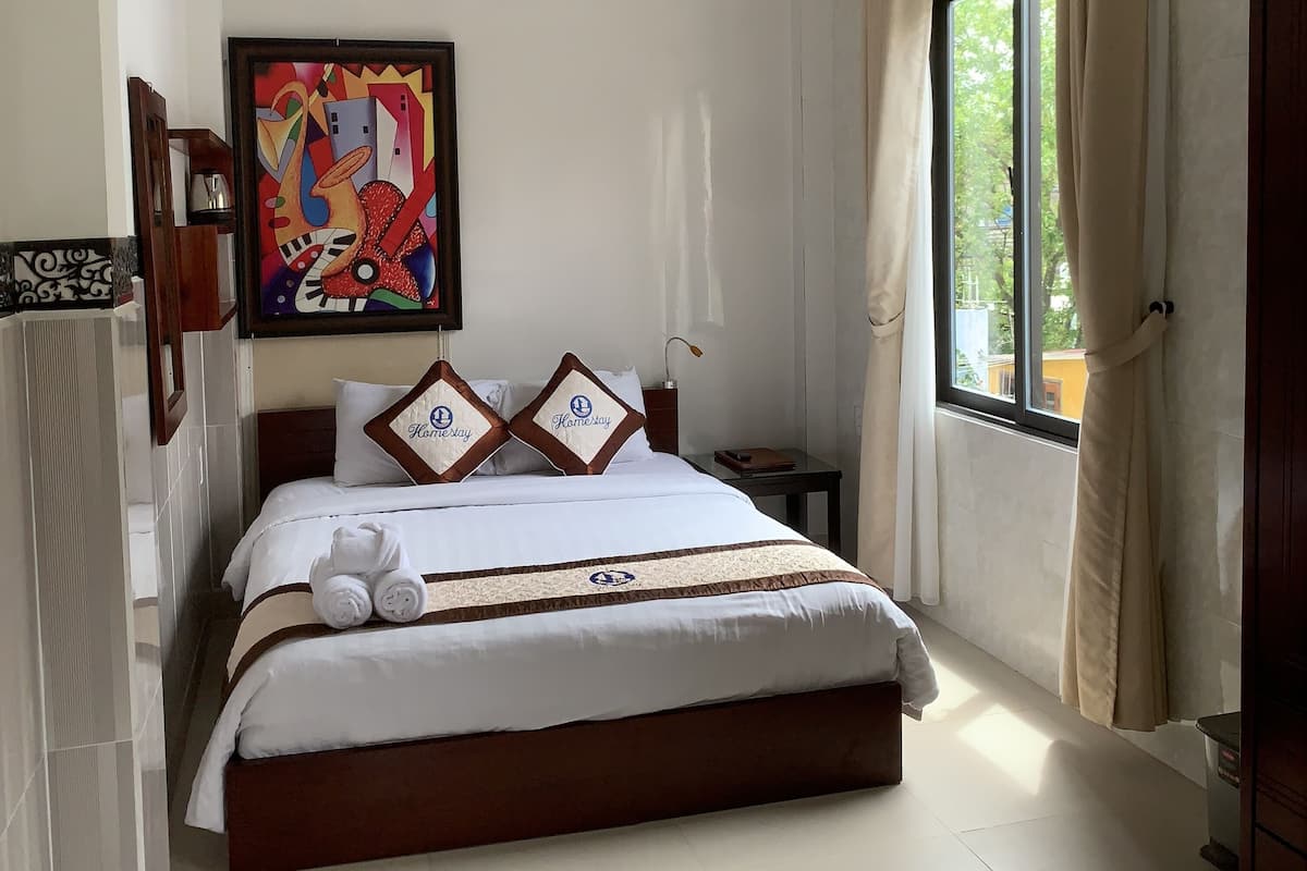 A hotel room with a bed, a ceiling fan, a window with curtains, and a painting on the wall.