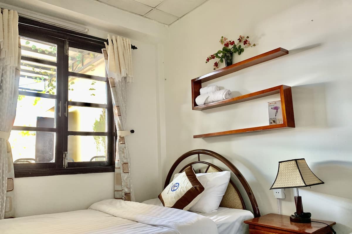 A hotel room with a bed, a nightstand with a lamp, a window with curtains, and a wall-mounted shelf with towels.