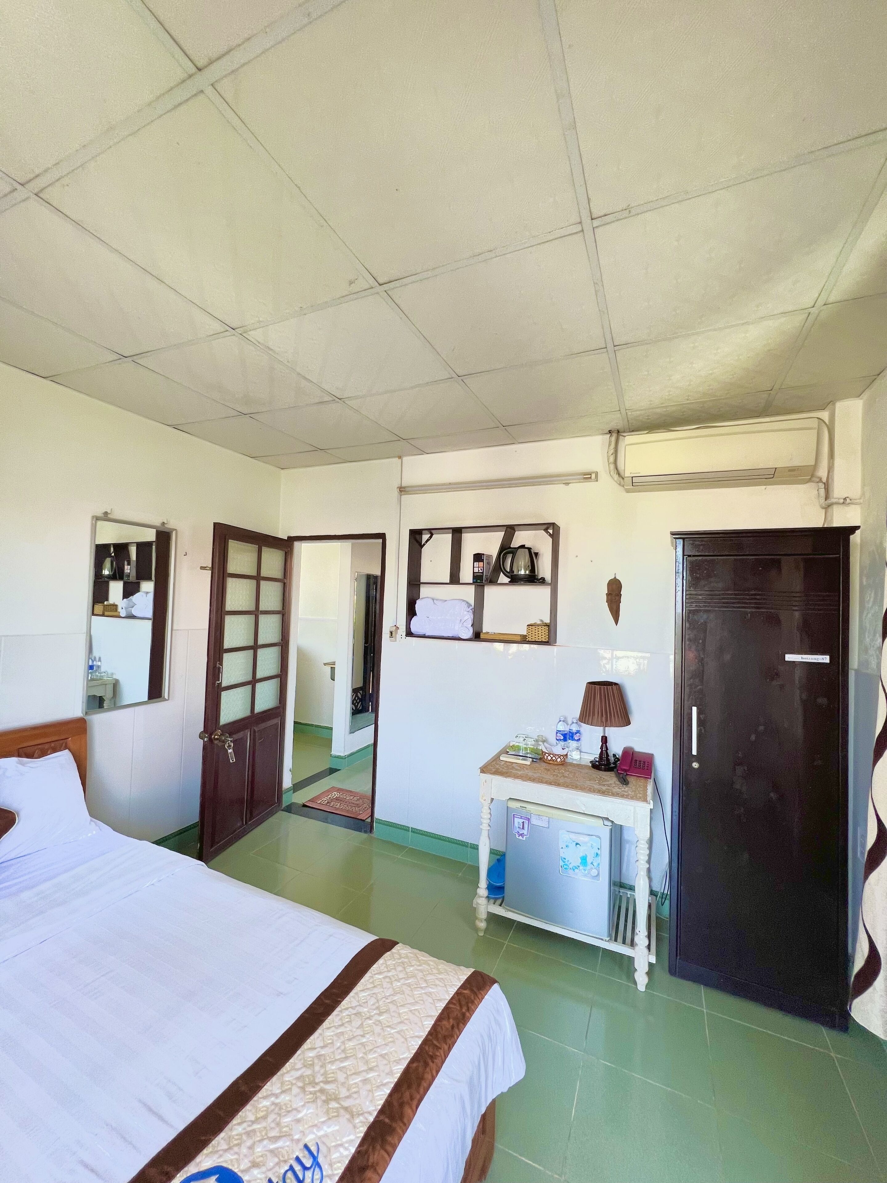 A hotel room with a bed, a desk, a mirror, a door, and an air conditioning unit.