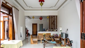 A living room with a sofa, coffee table, and a television. There are wooden chairs and a table with a yellow tablecloth. - Pink House Homestay (Da Nang)