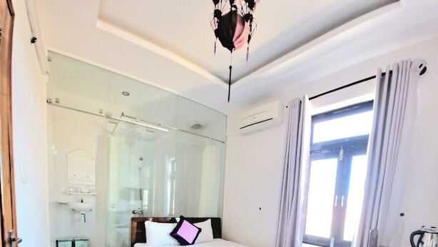 A hotel room with a large bed, a wooden desk, a glass shower enclosure, and a view of the city through the window. - Pink House Homestay (Da Nang)