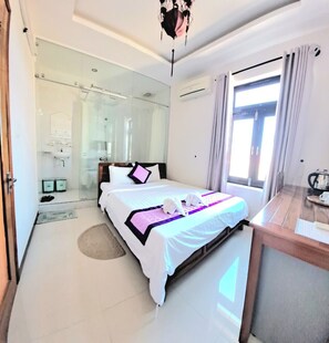 A hotel room with a large bed, a wooden desk, a glass shower enclosure, and a view of the city through the window. - Pink House Homestay (Da Nang)