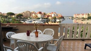 A terrace with a table and chairs, overlooking a marina with boats and buildings. - Spacious 3-bedroom apt with terrace, pool and parking near the beach (Port Barcarès)