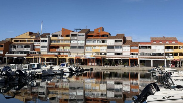 A marina with multiple boats docked, surrounded by multi-story buildings with balconies and various shops. - Spacious 3-bedroom apt with terrace, pool and parking near the beach (Port Barcarès)